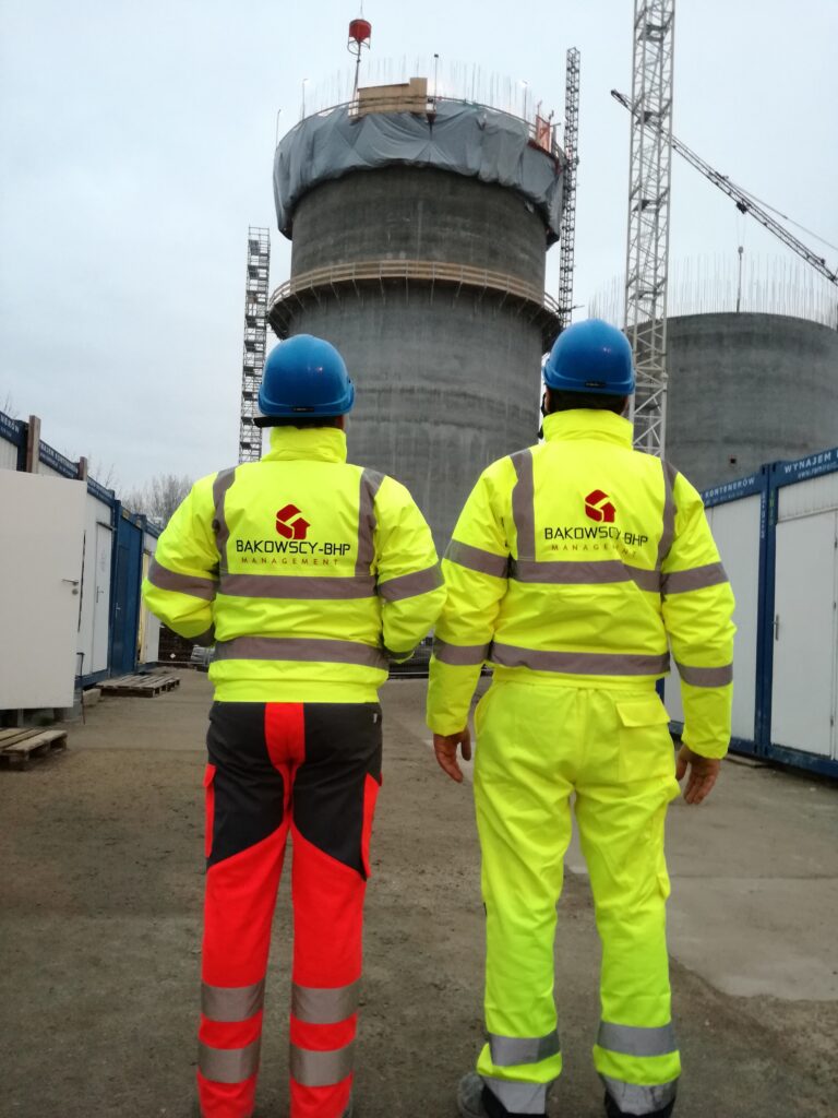 Dwie osoby w żółtych uniformach firmowych wraz z niebieskimi kaskami na budowie dwóch silosów podczas pełnienia obsługi BHP