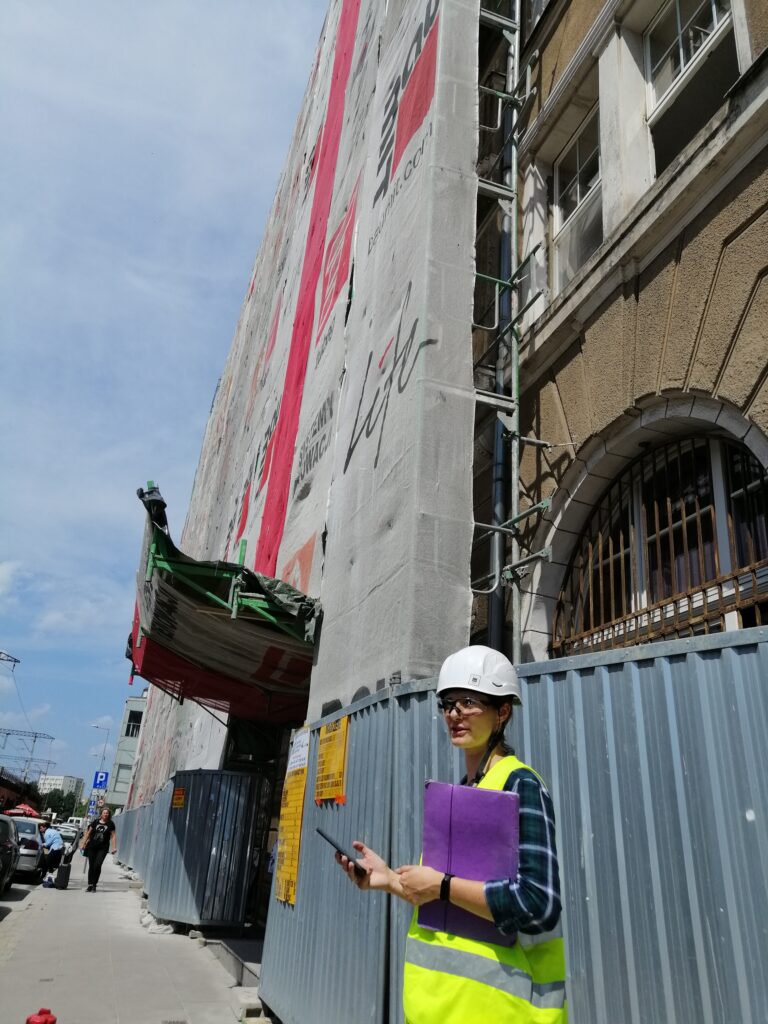 Kobieta stojąca w kasku BHP z żółtą kamizelką wraz z telefonem sprawdzająca stan robót budowlanych podczas nadzoru BHP